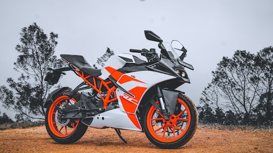 A sporty motorcycle with an angular design featuring a vibrant orange and white color scheme. It is parked on a dirt path surrounded by sparse vegetation and trees under an overcast sky, creating a contrast between the motorcycle's vivid colors and the muted background.