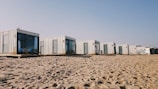 A row of modern, minimalist cabins made from shipping containers is positioned on a sandy beach. Each cabin features large windows and doors, some with outdoor seating. The sky is clear, suggesting a sunny day.