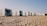 A row of modern, minimalist cabins made from shipping containers is positioned on a sandy beach. Each cabin features large windows and doors, some with outdoor seating. The sky is clear, suggesting a sunny day.