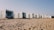 A row of modern, minimalist cabins made from shipping containers is positioned on a sandy beach. Each cabin features large windows and doors, some with outdoor seating. The sky is clear, suggesting a sunny day.