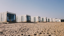 A row of modern, minimalist cabins made from shipping containers is positioned on a sandy beach. Each cabin features large windows and doors, some with outdoor seating. The sky is clear, suggesting a sunny day.