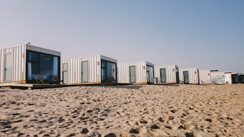 A row of modern, minimalist cabins made from shipping containers is positioned on a sandy beach. Each cabin features large windows and doors, some with outdoor seating. The sky is clear, suggesting a sunny day.