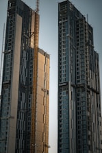 Two high-rise buildings under construction dominate the scene, featuring multiple floors with large glass windows. A yellow construction crane is situated between the buildings, indicating ongoing construction work. The structures have a modern design with a mixture of reflective panels and unfinished surfaces.