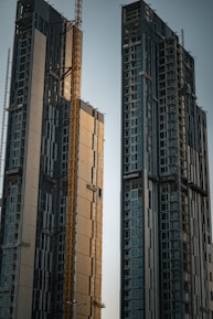 Two high-rise buildings under construction dominate the scene, featuring multiple floors with large glass windows. A yellow construction crane is situated between the buildings, indicating ongoing construction work. The structures have a modern design with a mixture of reflective panels and unfinished surfaces.