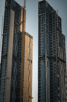 Two high-rise buildings under construction dominate the scene, featuring multiple floors with large glass windows. A yellow construction crane is situated between the buildings, indicating ongoing construction work. The structures have a modern design with a mixture of reflective panels and unfinished surfaces.