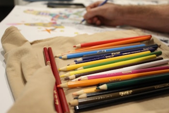 A variety of colored pencils are arranged neatly on a beige cloth, with a person in the background drawing on a piece of paper. The colored pencils are organized by color and are prominently placed in the foreground, while the person's hand is visible holding a blue pencil, working on a detailed artwork featuring various colors.