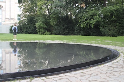A reflective circular pool surrounded by stone paving sits in a grassy area with trees and a building in the background. A person stands on the grass, looking towards the pool, wearing a backpack and casual clothes.