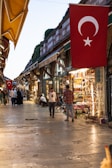 A bustling marketplace with narrow stone-paved pathways lined with various shops and stalls. People are walking and browsing the goods, which range from textiles to souvenirs. The area is illuminated with warm lights from the storefronts, and a large Turkish flag is prominently displayed overhead.