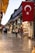 A bustling marketplace with narrow stone-paved pathways lined with various shops and stalls. People are walking and browsing the goods, which range from textiles to souvenirs. The area is illuminated with warm lights from the storefronts, and a large Turkish flag is prominently displayed overhead.