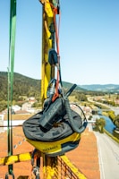 A toolbag labeled 'TOOLBAGS' is securely attached to a yellow, rusted metal structure with ropes and lanyards. The bag contains a radio or communication device with a coiled cord. The setting is high above a town with houses, roads, and a river, surrounded by green hilly landscape under a clear blue sky.