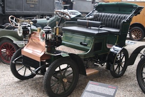 A vintage automobile with a classic open-top design featuring brass headlamps, a padded black leather bench seat, and green and black bodywork. The wheels have spoked rims and there's a copper-colored front section. Other antique vehicles are partially visible in the background.