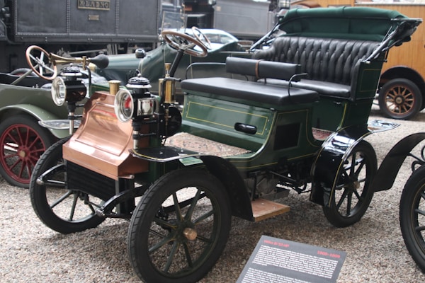A vintage automobile with a classic open-top design featuring brass headlamps, a padded black leather bench seat, and green and black bodywork. The wheels have spoked rims and there's a copper-colored front section. Other antique vehicles are partially visible in the background.