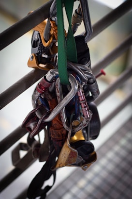 A collection of climbing equipment, including carabiners and belay devices, is hanging from a railing. The gear is attached to a green strap, showcasing several metallic items primarily in silver and gold tones.