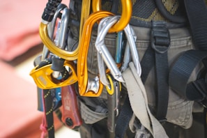 A climbing harness equipped with multiple carabiners and belay devices. The harness is gray, with bright orange and metallic tools attached, including a rope. The focus is on the gear's intricate details and sturdy design.