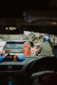 Two people are seen through a car windshield, with a child wearing a red jacket and hood holding a bouquet of flowers. Another person, wearing a blue helmet, appears to be interacting with the child. A car is parked in front, and the scene has a natural background.