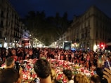 A large gathering of people surrounds a makeshift memorial with candles, flowers, and various tributes laid out. The setting appears to be a street in an urban area at night, with buildings lining the street and streetlights illuminating the scene. The crowd stands solemnly, suggesting a moment of reflection or mourning.