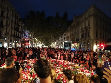A large gathering of people surrounds a makeshift memorial with candles, flowers, and various tributes laid out. The setting appears to be a street in an urban area at night, with buildings lining the street and streetlights illuminating the scene. The crowd stands solemnly, suggesting a moment of reflection or mourning.