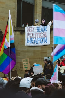 A protest scene with a crowd of people carrying signs and flags featuring LGBTQ+ colors. A prominent banner hanging from a building reads 'Not Your Political Weapon'. Several flags, including the rainbow and trans pride flags, are visible.