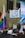 A protest scene with a crowd of people carrying signs and flags featuring LGBTQ+ colors. A prominent banner hanging from a building reads 'Not Your Political Weapon'. Several flags, including the rainbow and trans pride flags, are visible.
