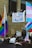 A protest scene with a crowd of people carrying signs and flags featuring LGBTQ+ colors. A prominent banner hanging from a building reads 'Not Your Political Weapon'. Several flags, including the rainbow and trans pride flags, are visible.