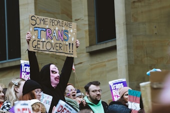 A group of people participating in a demonstration, with several holding signs advocating for transgender rights. The signs include messages of support and acceptance, such as a prominent one that reads 'Some people are trans, get over it!!' The crowd is diverse, and one individual stands out with a unique outfit that includes a pink knitted mask.
