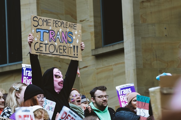 A group of people participating in a demonstration, with several holding signs advocating for transgender rights. The signs include messages of support and acceptance, such as a prominent one that reads 'Some people are trans, get over it!!' The crowd is diverse, and one individual stands out with a unique outfit that includes a pink knitted mask.