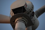 The close-up image captures the upper part of a white wind turbine, focusing on the nacelle and a section of the rotor blade. The turbine features various mechanical components and a metal rail around the nacelle.