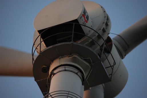 The close-up image captures the upper part of a white wind turbine, focusing on the nacelle and a section of the rotor blade. The turbine features various mechanical components and a metal rail around the nacelle.