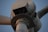 The close-up image captures the upper part of a white wind turbine, focusing on the nacelle and a section of the rotor blade. The turbine features various mechanical components and a metal rail around the nacelle.
