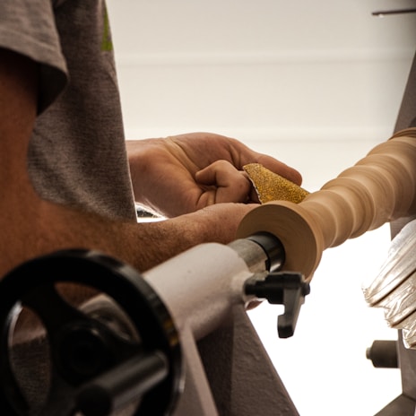 A person operates a lathe machine to shape a wooden object, holding sandpaper to smooth the surface. The image focuses on the craftsmanship involved in woodworking.