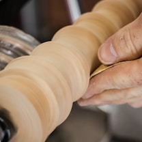 A close-up view of a hand holding sandpaper against a spinning wooden object, possibly on a lathe. The motion blur on the wood indicates it is rotating at a high speed.