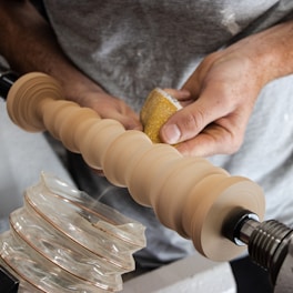 A person is engaged in woodturning, using a lathe to shape a cylindrical piece of wood. They are holding sandpaper, smoothing the surface with care. The lathe is in motion, and the wood has a light, natural hue.