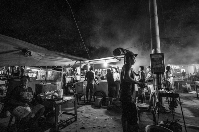 A bustling night market scene with people gathered around food stalls. The area is dimly lit, creating a moody and lively atmosphere. Various individuals are engaging in different activities, with some cooking and others conversing.
