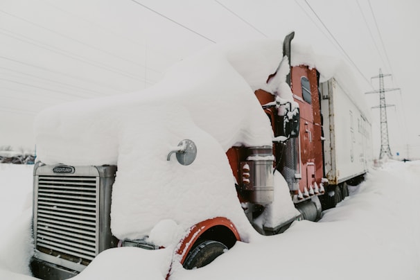 A large truck covered in a thick layer of snow is parked in a snowy landscape. The truck is mostly red with some visible chrome parts and a white cargo trailer. Snow is piled high on the truck's cab and hood, obscuring many of its features. In the background, there are power lines and a utility pole.