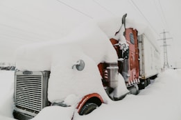 A large truck covered in a thick layer of snow is parked in a snowy landscape. The truck is mostly red with some visible chrome parts and a white cargo trailer. Snow is piled high on the truck's cab and hood, obscuring many of its features. In the background, there are power lines and a utility pole.
