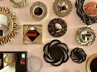 A collection of various decorative mirrors displayed on a wall, featuring intricate and diverse frame designs. Some frames are ornate and gold-colored with floral or geometric patterns, while others have a more modern and sleek black design. The reflections in the mirrors show parts of the surrounding environment and a couple of people.