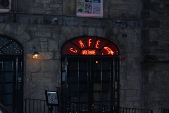 A dimly lit stone building features a café entrance with a prominent black double door. Above the door, a red neon sign reads 'CAFE VOLTAIRE' with a decorative lit outline. A warm, yellow light fixture is mounted on the wall to the left of the door. The atmosphere appears to be serene and cozy, with another square sign or menu stand visible near the entrance.