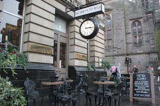 A street-side view of 'The Inn on the Mile' with its classic facade, featuring a large clock and traditional pub details. Outdoor seating with ornate wrought iron chairs and wooden tables is available. Several people are seen walking, and one person holds a Union Jack umbrella. A chalkboard sign advertises the pub's offerings.