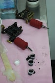 Tattoo equipment is arranged on a pink cloth atop a metal surface. There are two tattoo machines with red grips, small caps filled with black ink, a popsicle stick, and a plastic-wrapped item. A bottle of liquid is visible in the background.