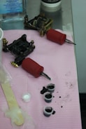 Tattoo equipment is arranged on a pink cloth atop a metal surface. There are two tattoo machines with red grips, small caps filled with black ink, a popsicle stick, and a plastic-wrapped item. A bottle of liquid is visible in the background.