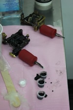 Tattoo equipment is arranged on a pink cloth atop a metal surface. There are two tattoo machines with red grips, small caps filled with black ink, a popsicle stick, and a plastic-wrapped item. A bottle of liquid is visible in the background.