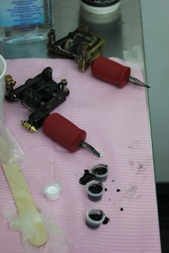Tattoo equipment is arranged on a pink cloth atop a metal surface. There are two tattoo machines with red grips, small caps filled with black ink, a popsicle stick, and a plastic-wrapped item. A bottle of liquid is visible in the background.