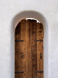 A rustic wooden door with visible grain patterns set within a rounded white plaster archway. The door features black metal hinges and a handle, creating a contrast between the warm wood tones and the bright white of the textured wall.