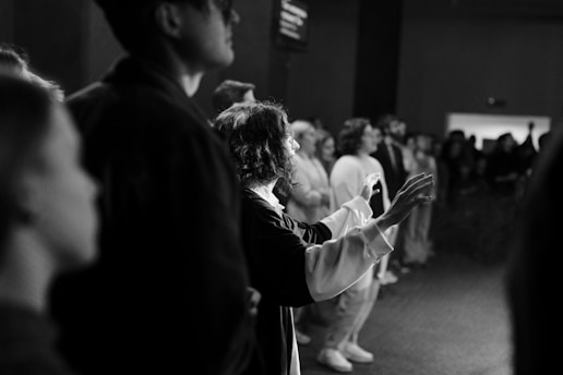 A black and white photograph depicts a group of people inside a dimly lit room. The focus is on an individual with curly hair and glasses, extending their hands, possibly in an expressive or thoughtful gesture. The surrounding crowd appears attentive or engaged in an event.