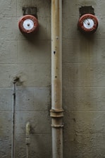 Two red fire alarm bells are mounted on a textured beige wall. A large, worn vertical pipe runs between them, and smaller, discolored pipes and rust marks are visible. The overall look is aged and industrial.