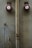 Two red fire alarm bells are mounted on a textured beige wall. A large, worn vertical pipe runs between them, and smaller, discolored pipes and rust marks are visible. The overall look is aged and industrial.