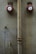 Two red fire alarm bells are mounted on a textured beige wall. A large, worn vertical pipe runs between them, and smaller, discolored pipes and rust marks are visible. The overall look is aged and industrial.