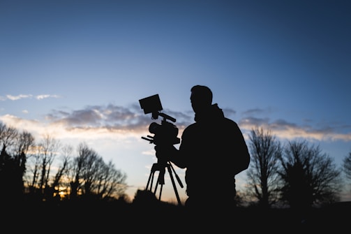 A silhouette of a person operating a video camera against a backdrop of a clear sky with scattered clouds and trees in the distance. The scene is captured during a twilight period, with the light dim yet distinct.