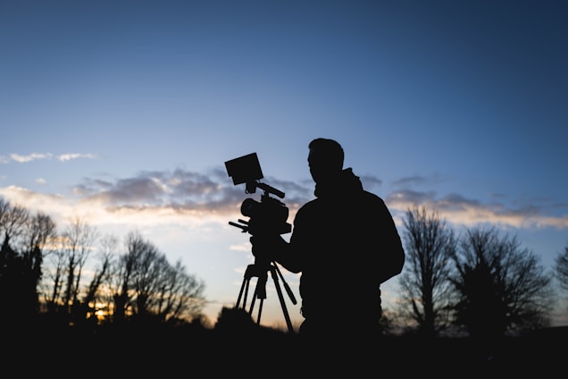 A silhouette of a person operating a video camera against a backdrop of a clear sky with scattered clouds and trees in the distance. The scene is captured during a twilight period, with the light dim yet distinct.