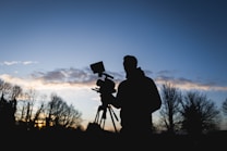 A silhouette of a person operating a video camera against a backdrop of a clear sky with scattered clouds and trees in the distance. The scene is captured during a twilight period, with the light dim yet distinct.